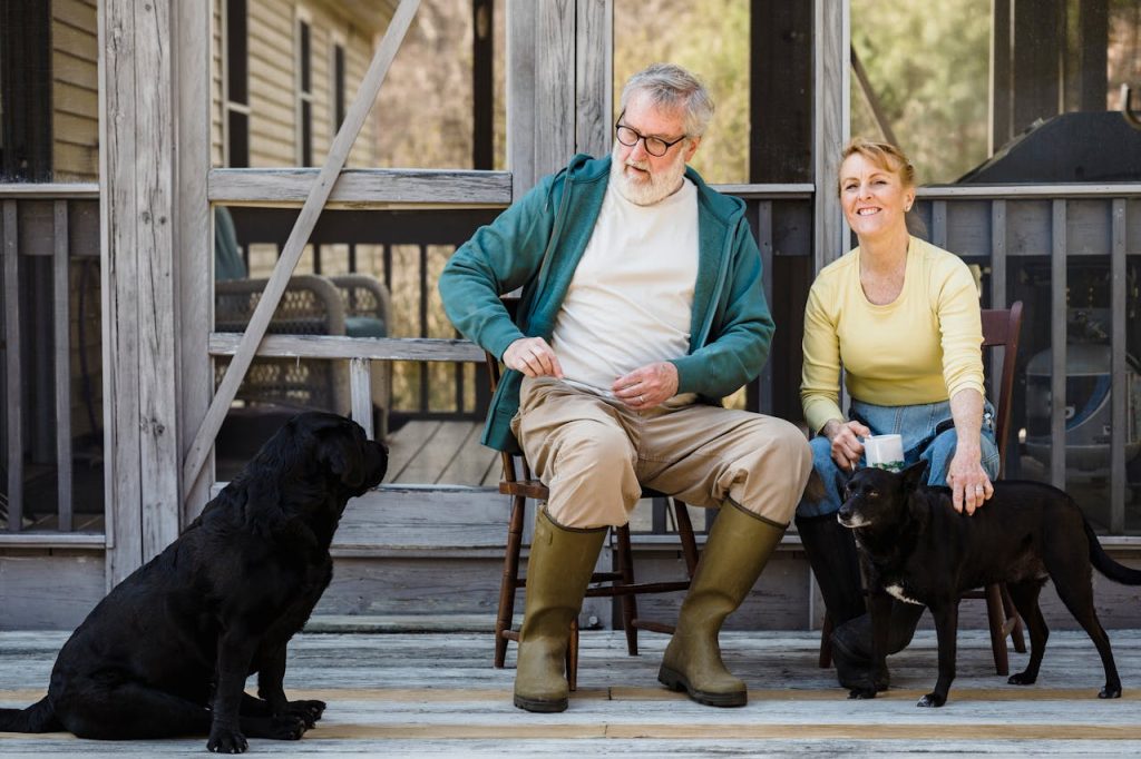 Delightful senior couple in rain boots sitting on chairs with black dogs while spending time together on terrace